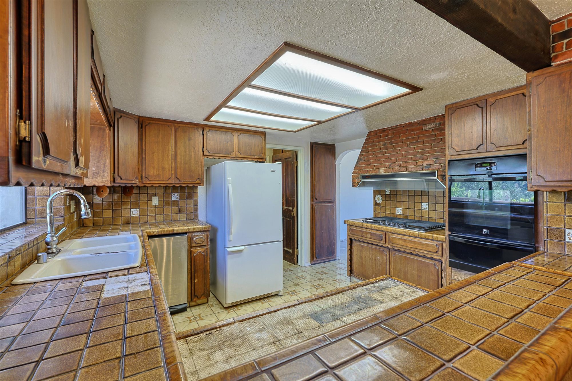 Existing kitchen with wood cabinetry, tile counters and a brick-faced hood surround